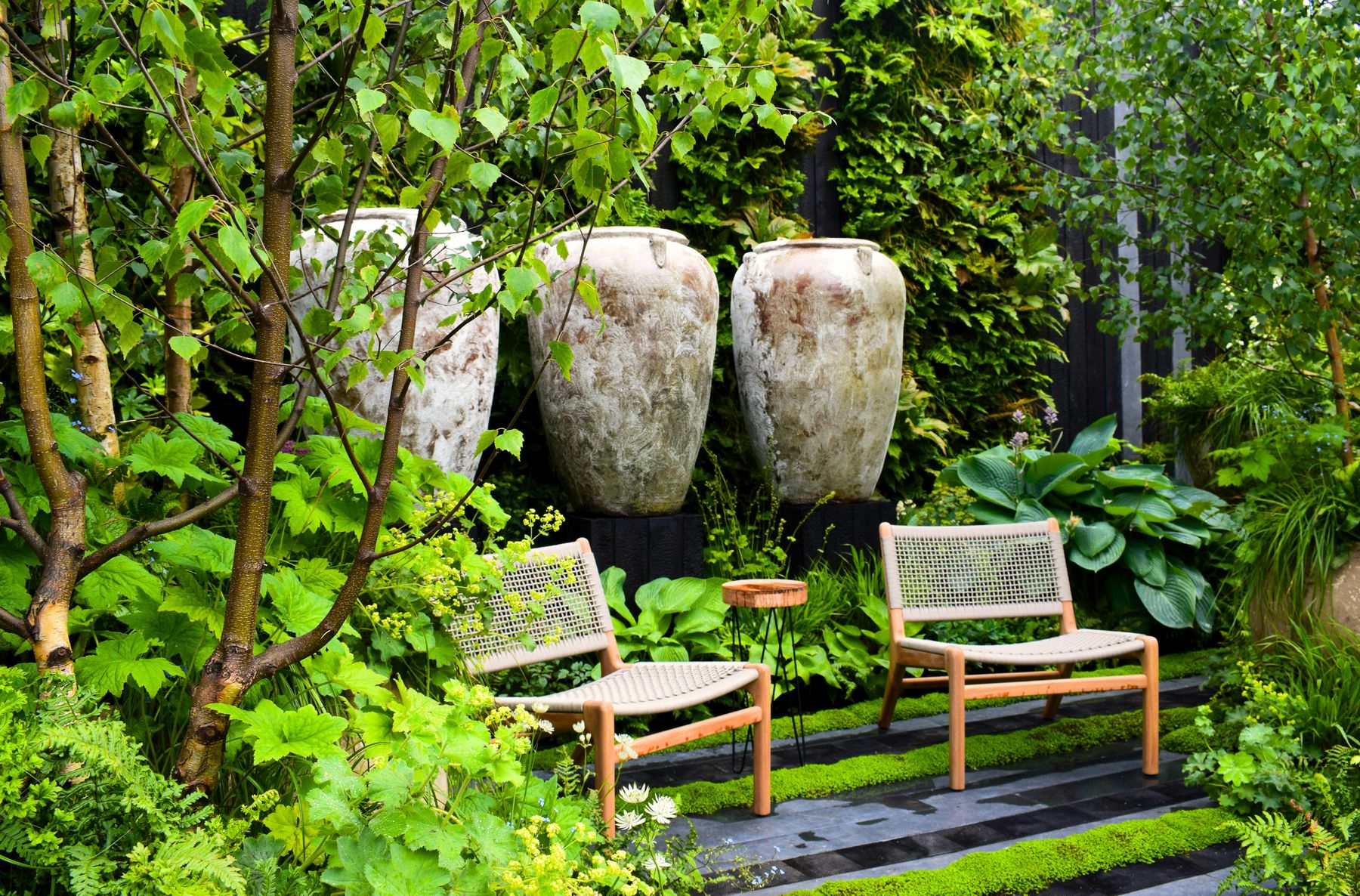 A lush established garden with aged stone urns, woven chairs and layered planting
